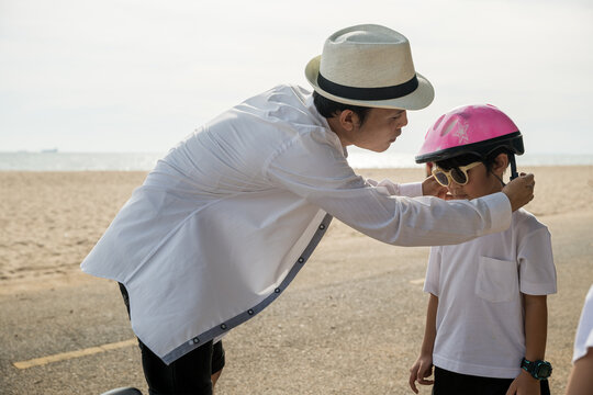 A family on vacation at the beach where the father sporting a safety helmet teaches his cheerful son the balance and joy of bicycle riding a memorable tourism day filled with family happiness.