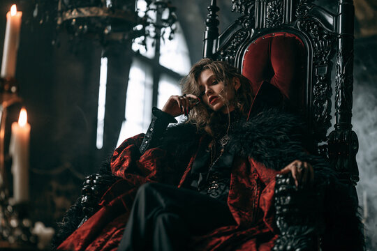 Aristocratic Vampire Woman Reclining on Ornate Gothic Throne in Luxurious Red and Black Halloween Costume by Candlelight