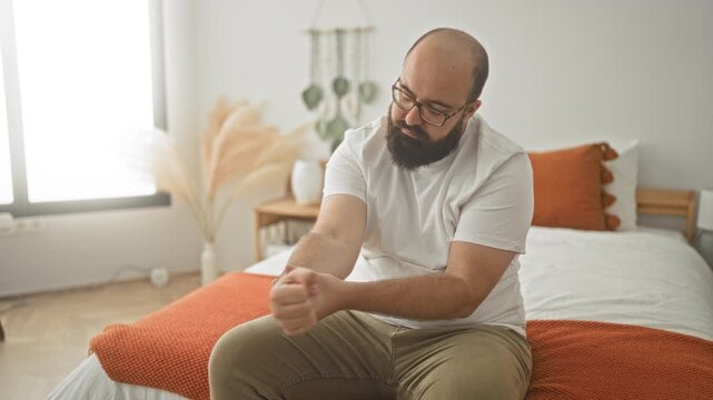 Man massaging wrist and rubbing forearm while seated on bed in bedroom with orange blanket and pillow; pain recovery healing.