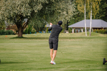 Fototapeta premium Golfer in dark clothing completing a full swing across manicured fairway, viewed from behind with trees and clubhouse in the background at an outdoor golf course