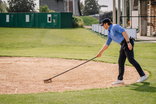 Golfer in blue shirt and cap raking sand bunker with long handled tool beside manicured fairway and clubhouse buildings at outdoor golf course