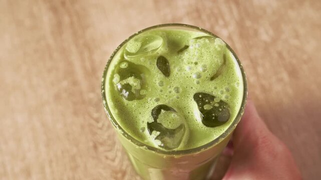 Cold iced green matcha latte with ice cubes and foam inside clear glass on wooden table. Refreshing summer antioxidant detox superfood matcha beverage for healthy daily routine.