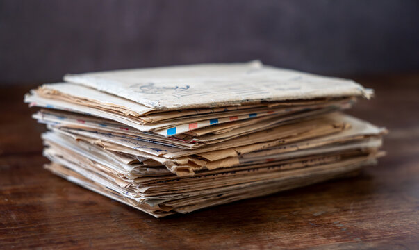 A stack of old letters rests on a wooden table. Frayed envelopes and handwritten pages show layered paper. Archival correspondence evokes memory, history, and analog recordkeeping.