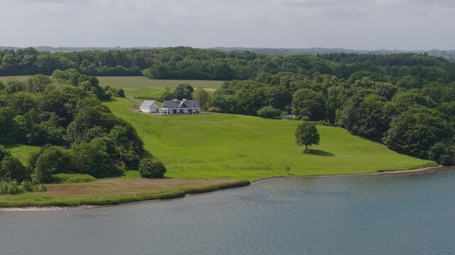 Drone footage showcasing a secluded cottage nestled on a green hillside near Kolding Fjord, with scenic woodland, meadows, and calm fjord waters in southern Denmark.