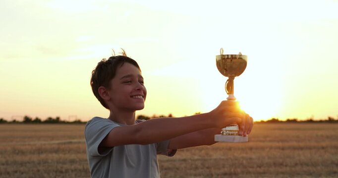 Happy boy holds trophy in golden wheat field at sunset. Young winner celebrates sports achievement outdoors. Success, pride and victory inspire dreams, motivation and childhood joy after competition.