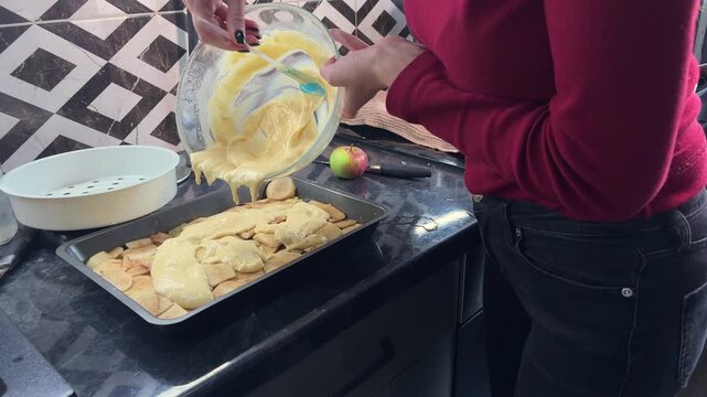 Close up of woman pouring smooth batter over apple slices in a baking dish, homemade cake preparation