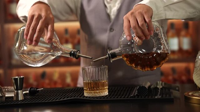 Professional bartender pours clear spirit and dark whiskey from glass decanters into a rocks glass at a warm bar counter.