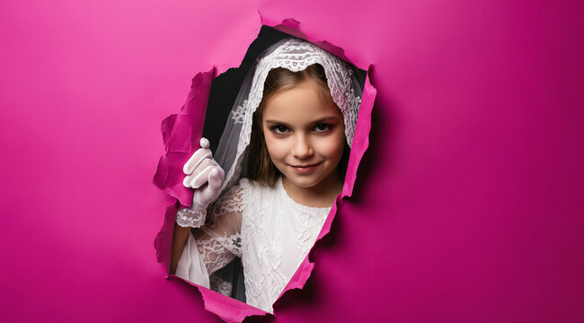 Young girl peeking through a torn hole in pink paper, wearing white lace veil and gloves. Playful expression, creative concept. Childhood, fashion, costume, celebration.