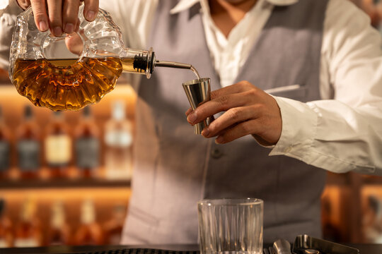 Bartender pours amber whiskey from a crystal decanter into a metal jigger at a warm, softly lit bar.
