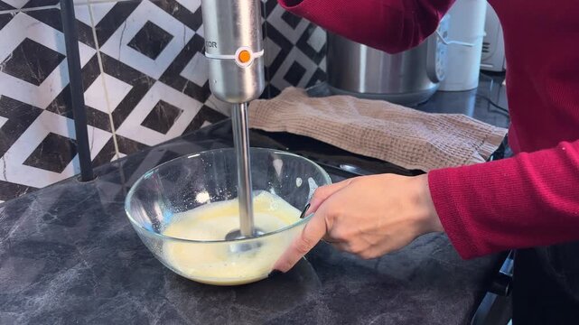 Woman blends eggs and sugar with an immersion blender in a glass bowl, making batter for a homemade dessert