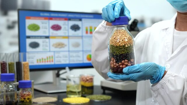 Food researcher in a laboratory holds a glass jar of layered beans, lentils and grains beside a computer screen showing sample data.