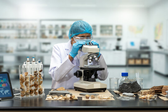 Focused laboratory scientist in protective clothing using a microscope to examine natural samples, with test tubes, rocks, wood pieces and a data tablet on the workbench.