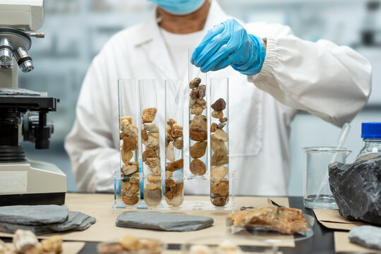 Scientist in protective gloves places rock samples into glass test tubes beside a microscope in a laboratory, studying minerals and geology.
