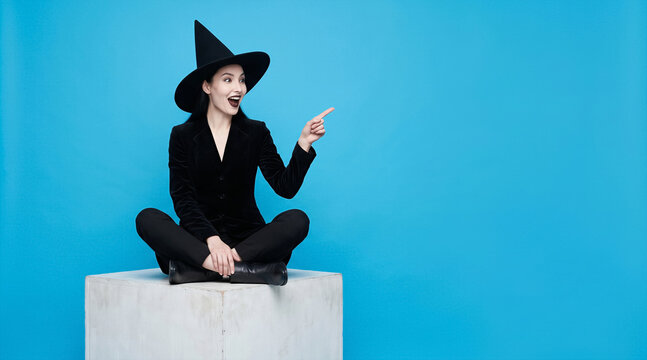 Young woman in witch costume sitting on white cube and pointing to side, surprised expression, blue background, copy space. Halloween, costume, celebration, party idea.