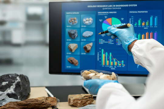 Geology researcher in blue gloves examines rock samples while pointing at a digital dashboard with mineral images, charts and graphs in a modern laboratory.