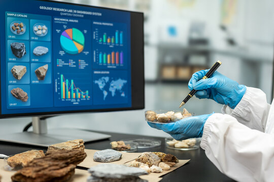 Geologist in gloves examines rock samples with a pen beside a monitor showing mineral data charts and maps in a modern laboratory.