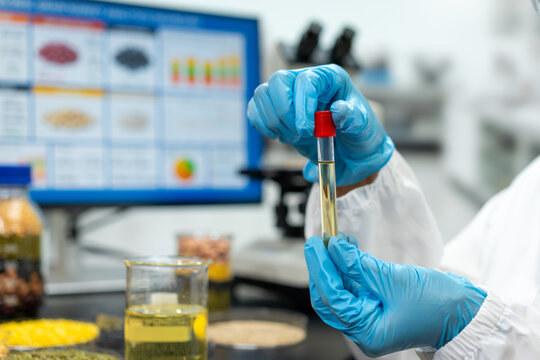 Scientist wearing blue gloves holds a test tube with yellow liquid in a laboratory, with a computer monitor, microscope and glass beaker in the background.
