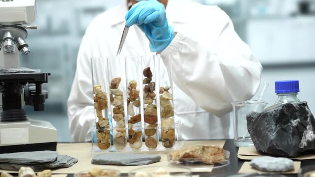 Scientist in a white lab coat uses tweezers to examine rock samples in glass test tubes beside a microscope in a modern laboratory.