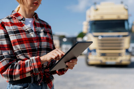 A female logistics professional checks data on a digital tablet with a large commercial vehicle parked behind her on a sunny day