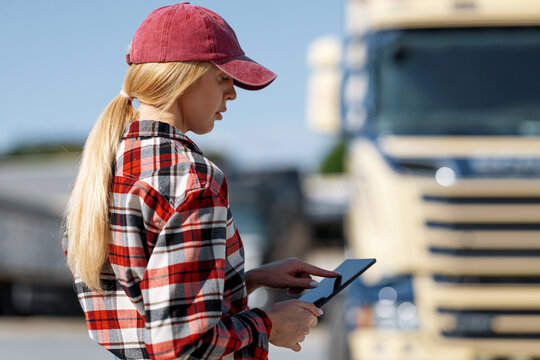 A female trucker in a red cap and plaid shirt checks her digital device while standing beside a large commercial vehicle outdoors