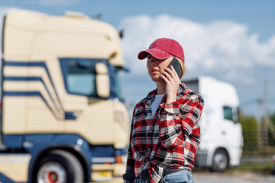 A female trucker wearing a red cap and plaid shirt is on a phone call while standing outdoors with large transport vehicles in the background