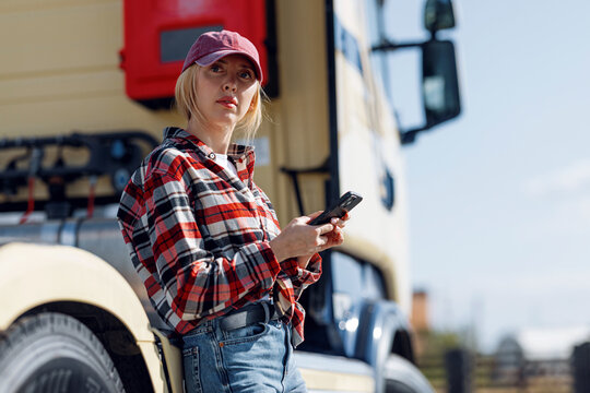 A female truck driver in a plaid shirt and baseball cap checks her phone while standing next to her large commercial vehicle on a sunny day