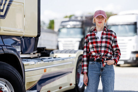 A confident woman wearing a red and black plaid shirt and a pink baseball cap poses in front of a fleet of large transport vehicles