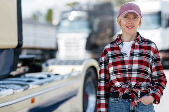 A confident woman wearing a red plaid shirt and a baseball cap poses in front of a large semi-trailer truck on a sunny day
