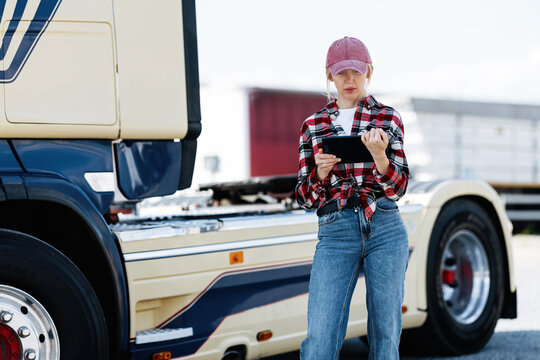A female trucker in a plaid shirt and baseball cap checks her digital device while standing beside a large commercial vehicle