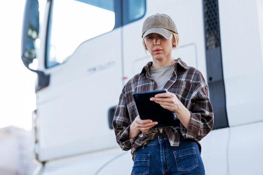 The woman wears a plaid shirt and baseball cap while checking logistics or delivery schedules on her mobile device during a stop at the depot