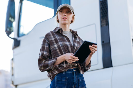 The professional driver checks her digital device while standing outdoors near the side of a heavy freight vehicle during a bright sunny day
