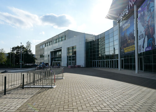 Modern exhibition center in Cologne exterior with glass facade, promotional gaming banners, paved forecourt, entrance doors, and temporary crowd barriers at gamescom trade fair venue