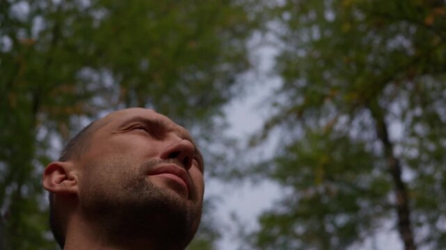A man with a short buzz cut and light stubble gazes upward with eyes slightly closed, head tilted back against blurred green foliage and a pale sky, while small insects fly past his face.