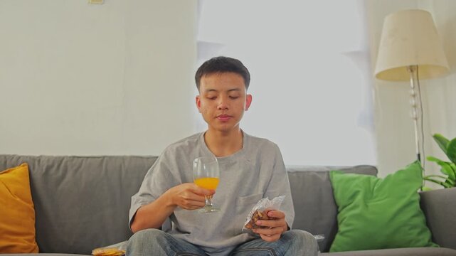 Young man drinking juice and broad beans while sit on cozy sofa