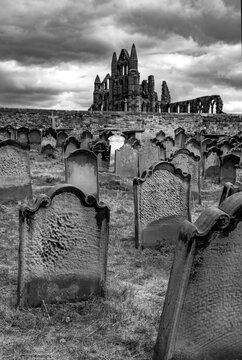 Weathered gravestones in the historic churchyard of St Mary&rsquo;s Church with the ruins of Whitby Abbey rising behind them beneath dramatic storm clouds, Whitby. Black and white gothic atmosphere.