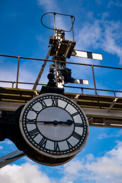 Vintage railway clock with Roman numerals beneath traditional semaphore signals at Grosmont Station, North Yorkshire, against a vivid blue summer sky.