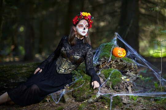 Halloween beauty portrait of young woman in the forest on a mossy stump with a halloween pumpkin.