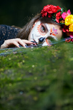 Halloween beauty portrait of young female with Santa Muerte makeup lies on tree trunk in the forest