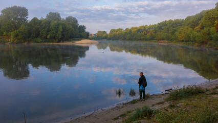 A person stands on the misty riverbank, reflecting the serene morning sky and lush green trees. © Александр Арендарь