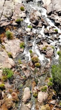 Vertical aerial view of rocky creek channel with flowing water, foamy cascades, and dense bank vegetation over fractured bedrock and scattered boulders in bright sunlight