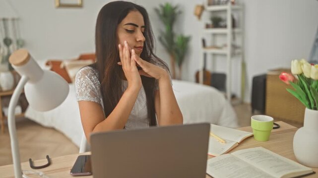 Arab woman using laptop with hands on cheeks at desk in building with books and lamp visible; boredom remote work.