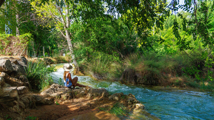 Woman wearing hat relaxing by river bank surrounded by green forest trees during summer nature travel