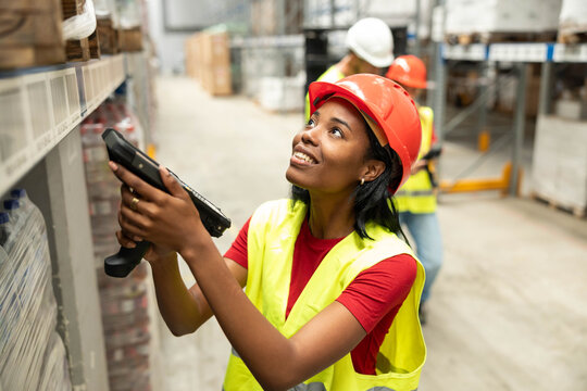 Woman in hard hat and safety vest scanning inventory on high shelves with a barcode reader in a busy logistics warehouse