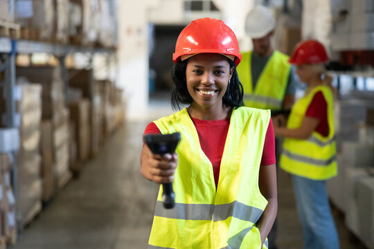 Smiling african american woman working in a warehouse, wearing safety vest and hard hat, scanning inventory with a barcode reader
