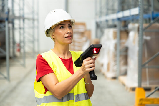 Female warehouse worker wearing hard hat and safety vest, scanning inventory with a barcode reader in a modern distribution center