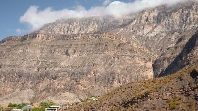 Massive layered mountain cliffs with low cloud over dry foothills and small hillside settlement under clear blue sky, showing serene remote highland landscape, rugged stone texture, and expansive