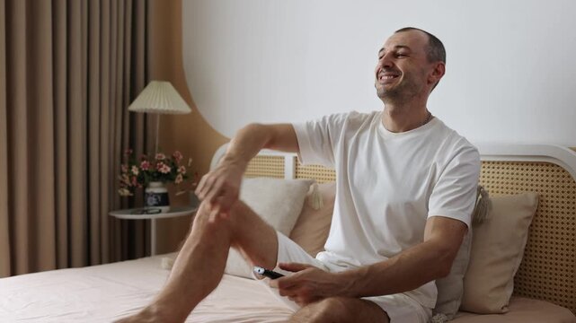 Young man sitting on bed, holds remote control, watching TV and smiling.