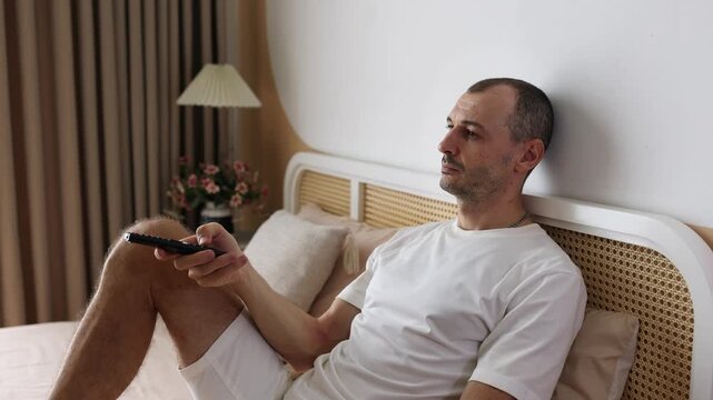 Young man sitting on bed, watching TV. He is bored and just switching channels.