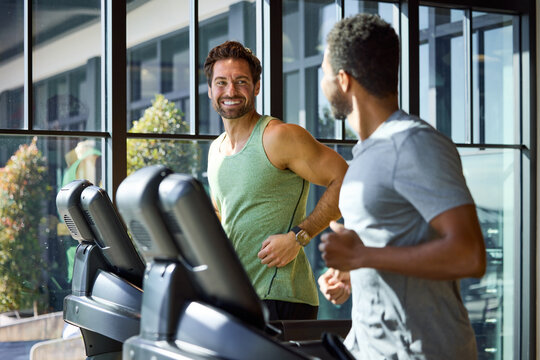 Two men jogging on treadmills in a bright gym. They smile and chat while exercising, enjoying their workout. Office in-house gym.