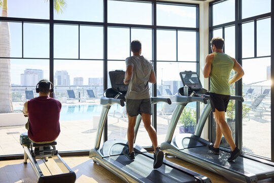 Three people exercising on treadmills and rowing machine with a city view. Cityscape view. Office in-house gym. From behind